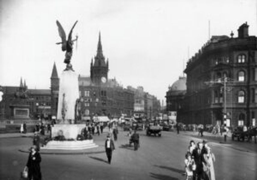 Black-and-white historic photograph of a busy city square with a large stone war memorial topped by a winged statue, surrounded by pedestrians and early motor vehicles, with grand buildings and a church tower in the background.