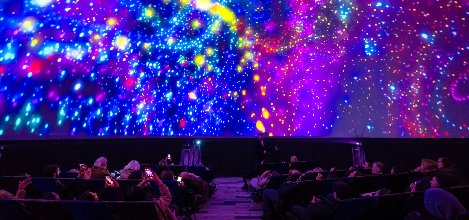 Visitors watch a colourful immersive galaxy projection on the dome screen of a planetarium.