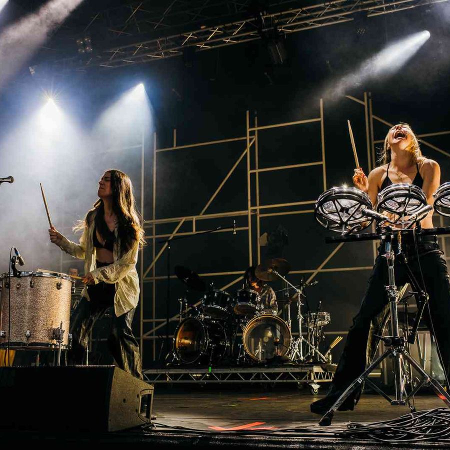 Two members of Haim performing with percussion instruments and drum kit on lit stage.
