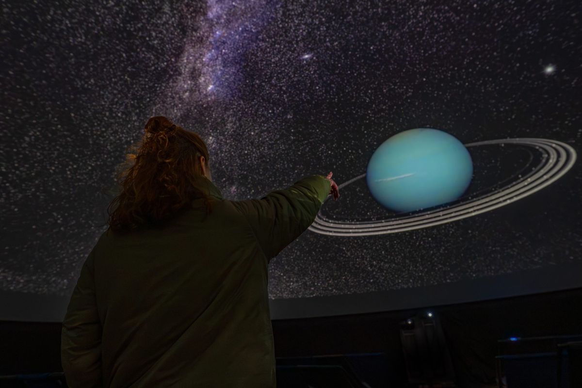 Visitor points towards a projected image of a ringed planet inside an immersive planetarium dome.