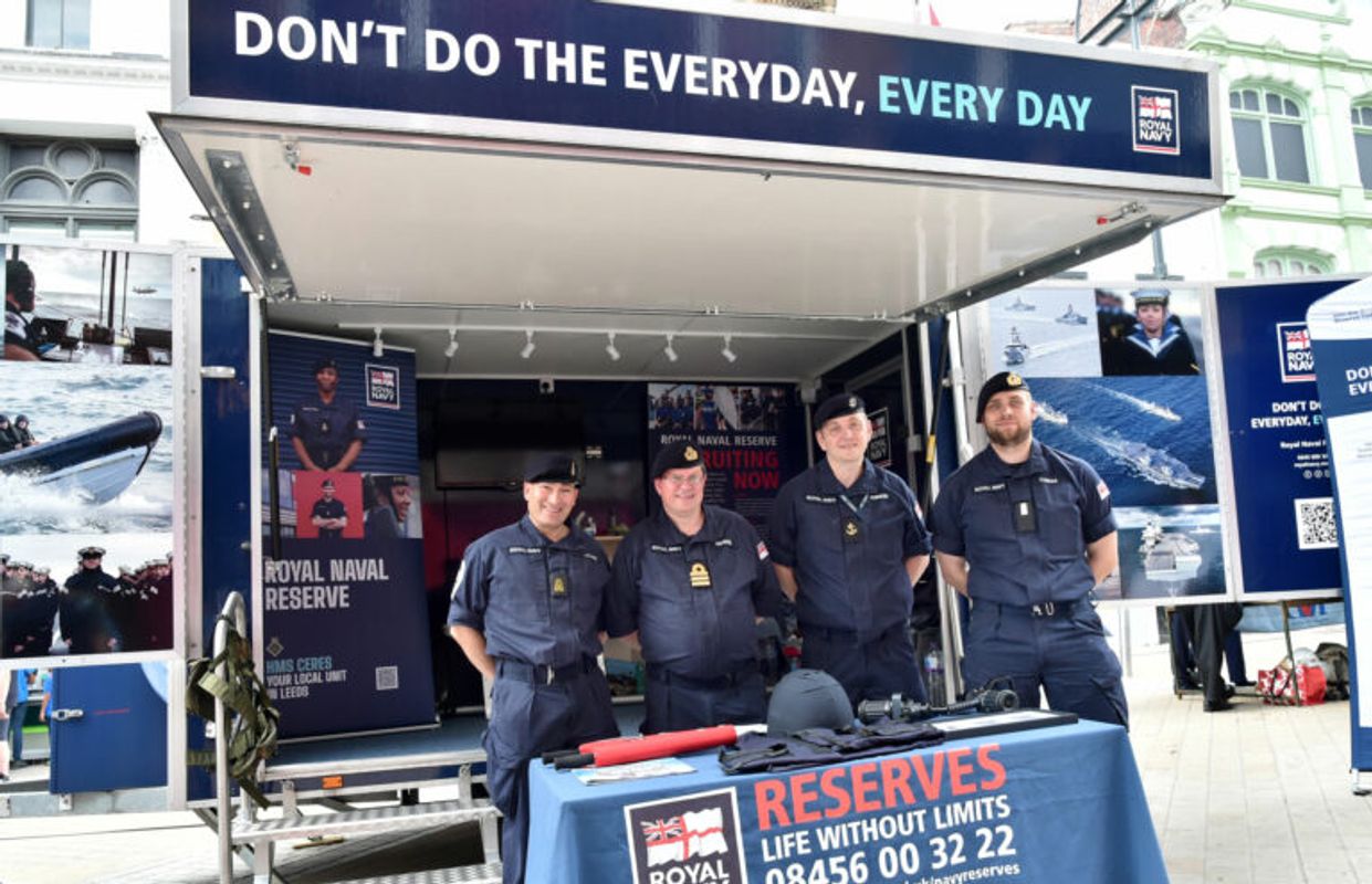 Four Royal Naval Reserve personnel stand behind a recruitment display table outside a mobile exhibition unit,
