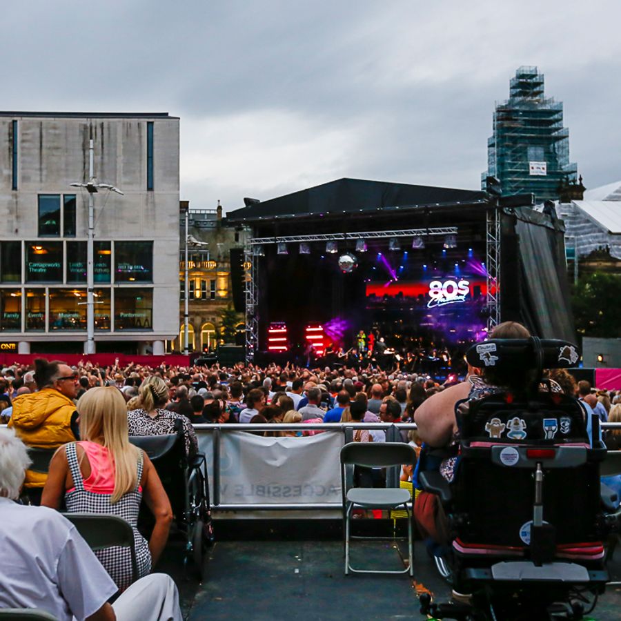 Outdoor city-centre concert at dusk with a large crowd facing a lit “80s Classical” stage, accessible seating in the foreground including a mobility scooter user, and surrounding buildings under a cloudy sky.