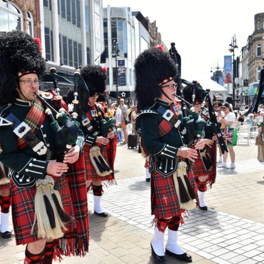 A marching group of bagpipe players in traditional tartan kilts and tall black feathered hats perform on a busy pedestrian street lined with shops and spectators.