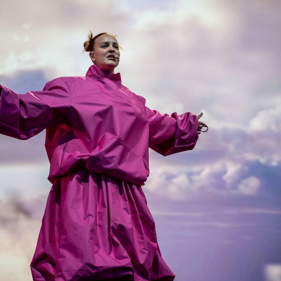 Anne-Marie in a bright pink oversized outfit on stage at Millennium Square, standing with arms outstretched against a soft cloud backdrop.