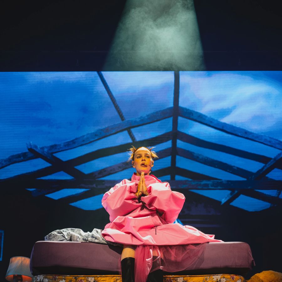 Anne-Marie in a bright pink outfit seated on stacked bed props on stage at Millennium Square, under cool blue lighting with a spotlight overhead.