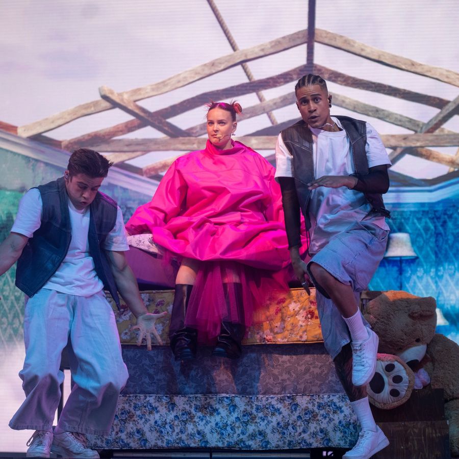 Anne-Marie in a bright pink outfit seated on a bed prop on stage at Millennium Square, performing alongside two dancers within a bedroom-style set.