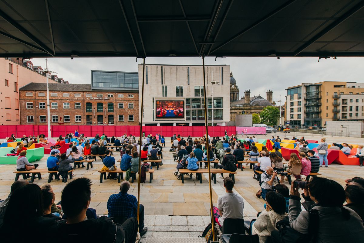 Crowd seated on benches in Millennium Square Leeds watching content on Leeds Big Screen outside the Carriageworks Theatre