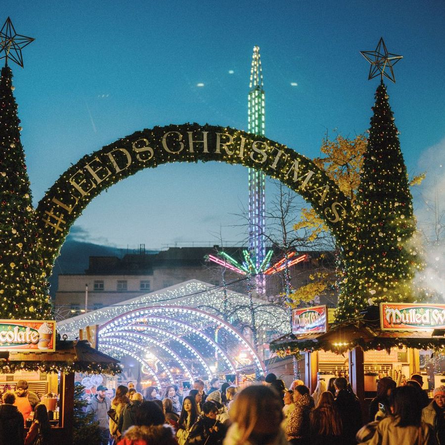 Crowds gathered around festive food and drink stalls beneath illuminated Christmas trees and the Leeds Christmas arch, with seasonal attractions and festive lighting in the background.