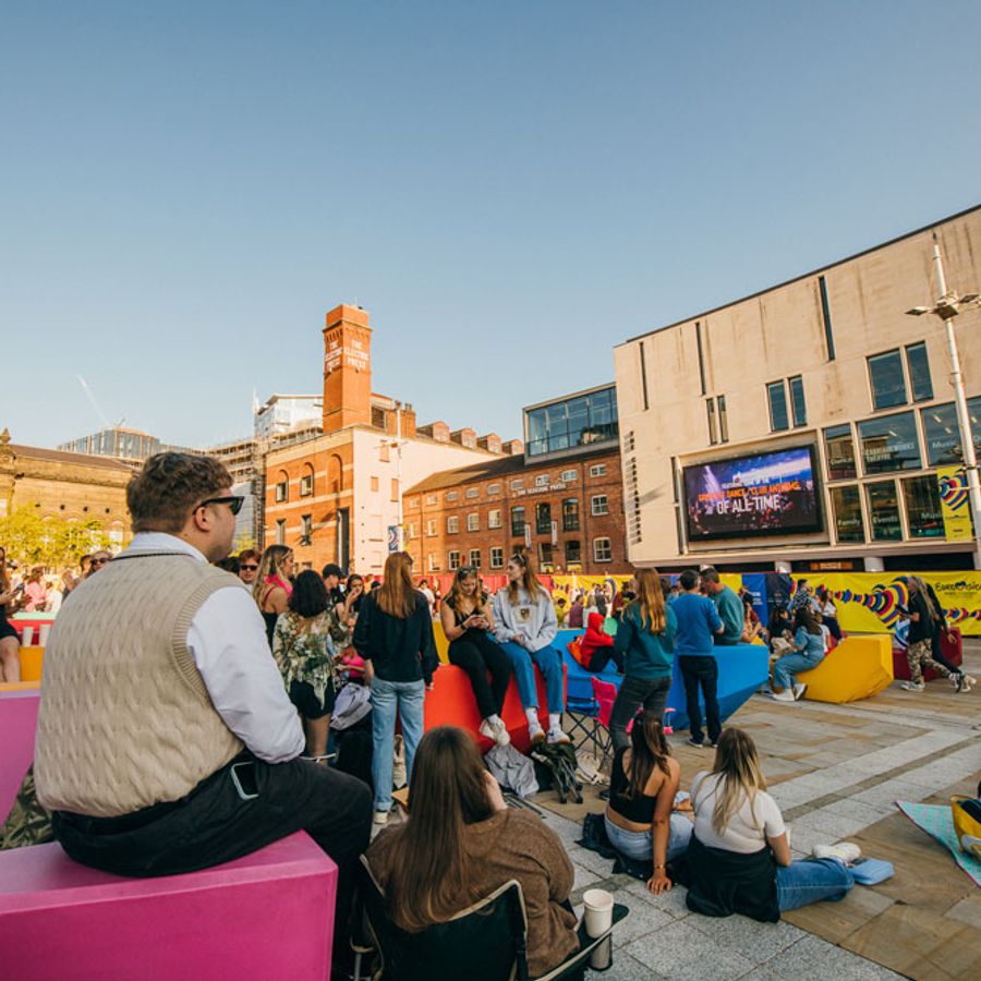 People sitting on colourful benches watching content on the Leeds Big Screen outside the Electric Press building on a sunny day