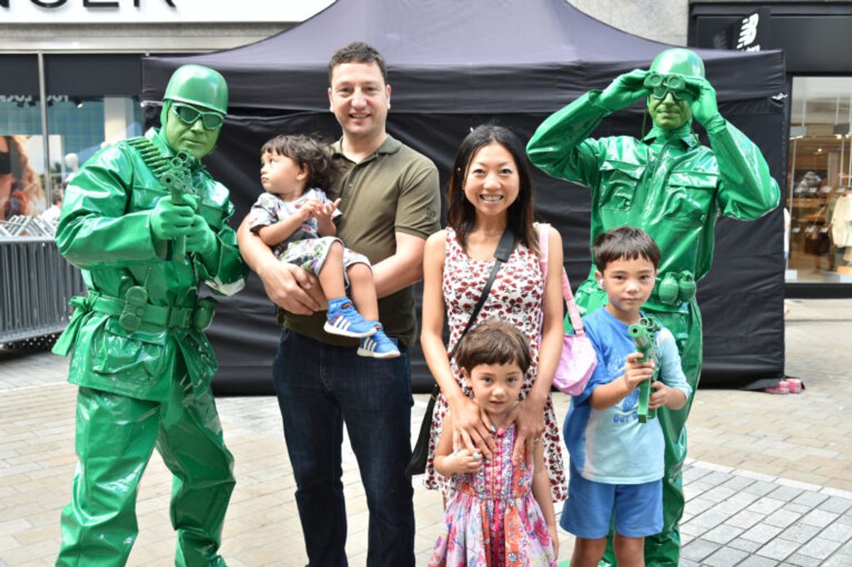 A family poses with two performers dressed as green toy soldiers, who are holding plastic toy guns and standing in front of a black event tent on a pedestrian street.