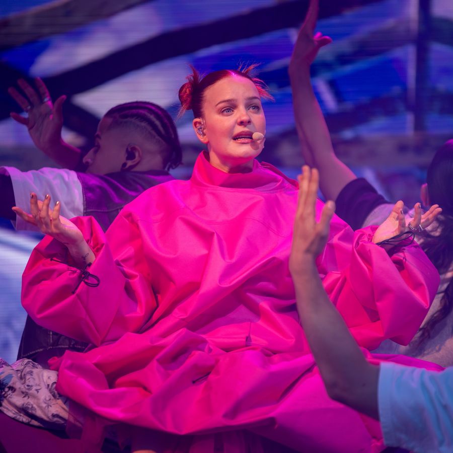 Anne-Marie in a bright pink oversized outfit on stage at Millennium Square, standing with arms outstretched against a soft cloud backdrop.