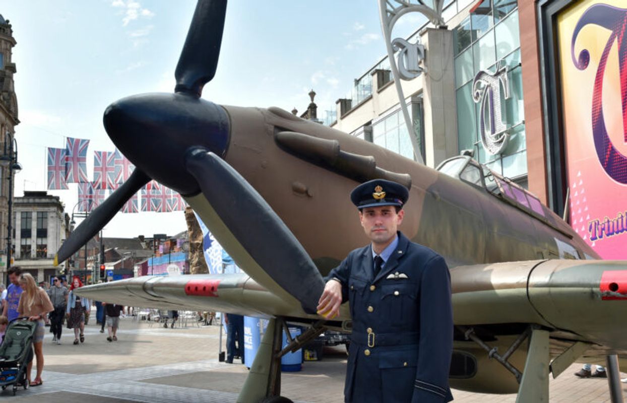 A person in a dark blue military-style uniform stands beside the propeller of a vintage aircraft displayed in a public square decorated with Union Jack flags.