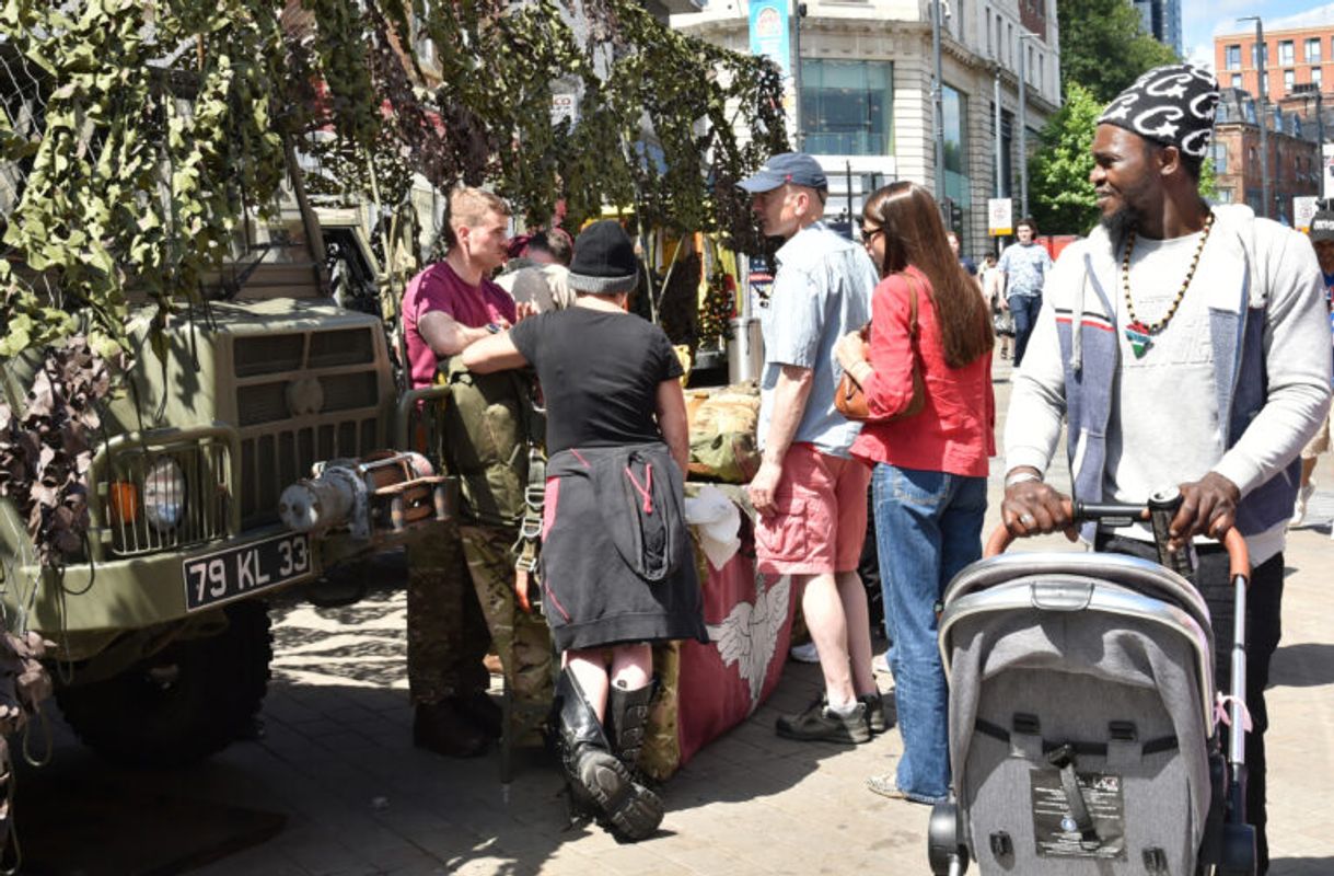 People gather around a military display vehicle draped in camouflage netting while a person pushes a stroller past the scene on a sunny city street.
