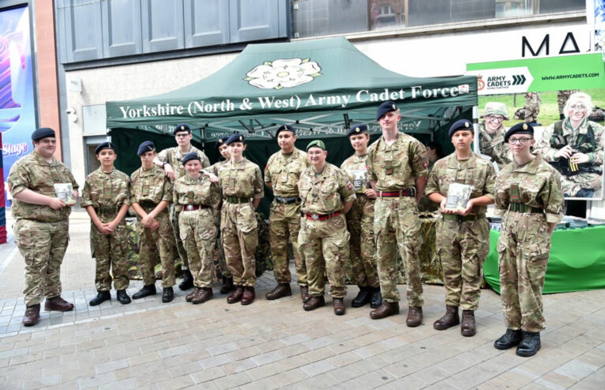 A group of army cadets in camouflage uniforms stand in a line in front of a green tent labelled “Yorkshire (North & West) Army Cadet Force,” holding leaflets on a city street.