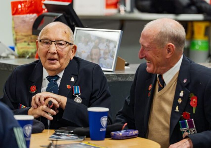 Veterans wearing medals sit around a table in a café setting, talking together with cups and leaflets on the table.