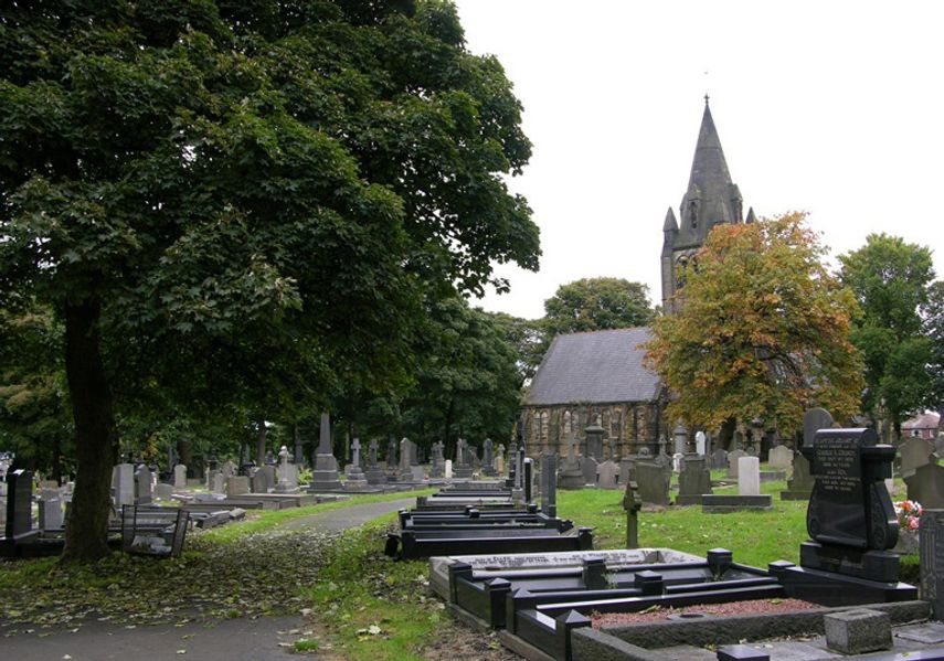 A quiet cemetery with rows of gravestones and a stone church in the background, surrounded by trees and greenery.