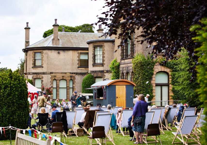 People relax on deck chairs and gather in the garden of Lotherton Hall during an outdoor event, with bunting, a small wooden hut and greenery surrounding the scene.