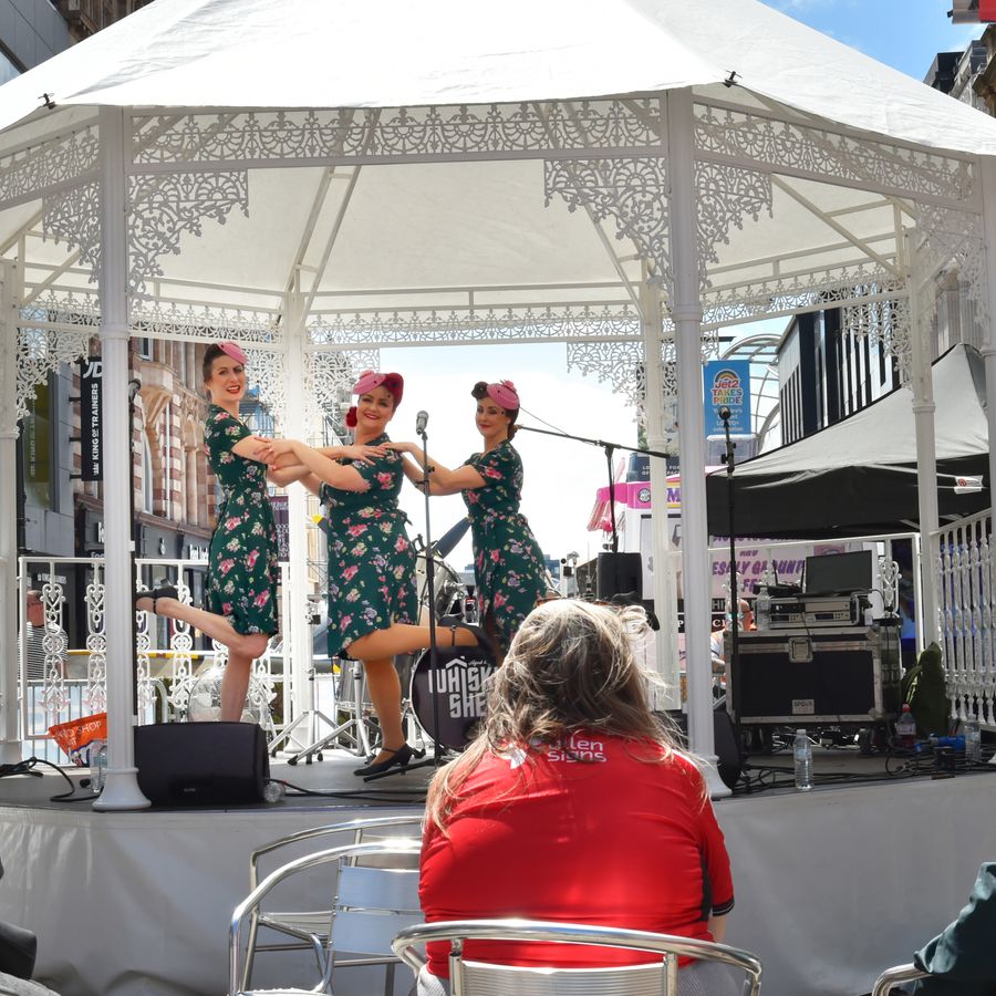 Alt text: Three performers in 1940s-style floral dresses and hats pose mid-routine on a small outdoor stage under a white decorative gazebo on a busy city street, with an audience seated in front and shopfronts in the background.