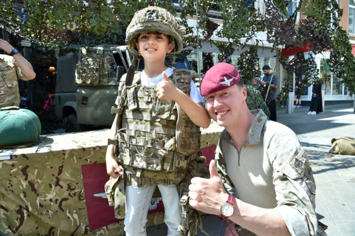 A child wearing camouflage military-style clothing stands beside an adult in uniform with a red beret, both giving thumbs up in front of a display vehicle covered in netting.