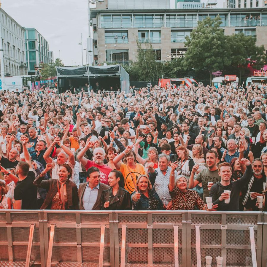 Crowd of people standing on Millennium square smiling and with their hands raised in the air.