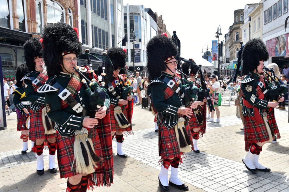 A marching group of bagpipe players in traditional tartan kilts and tall black feathered hats perform on a busy pedestrian street lined with shops and spectators.