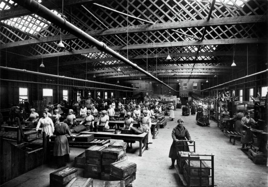 A black-and-white photograph of a busy wartime factory interior, showing rows of workers assembling munitions at long tables under a high industrial roof.
