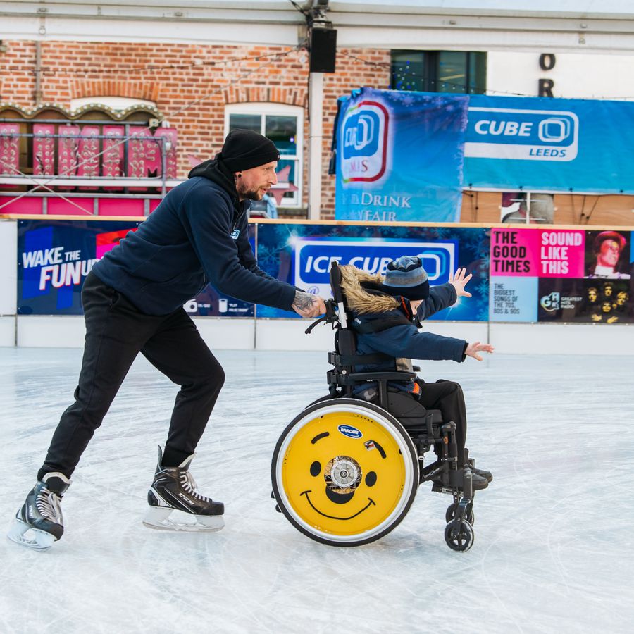 Person on ice skates pushes a wheelchair user across an outdoor ice rink in Leeds.
