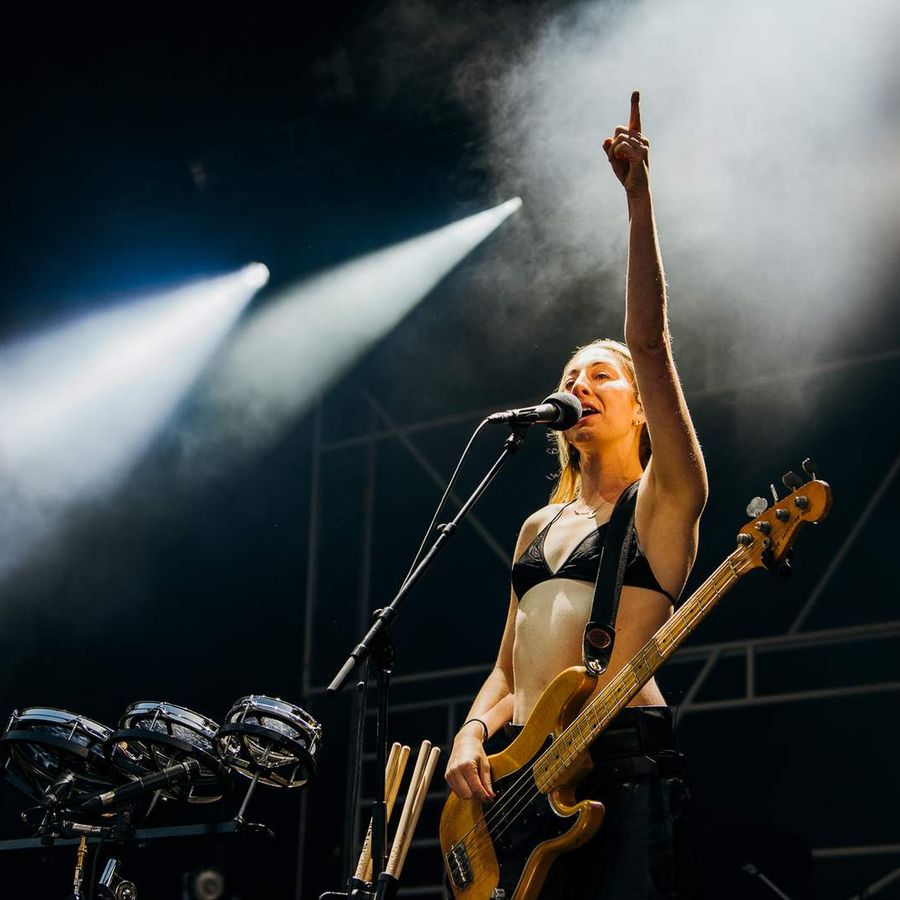 Musician playing bass guitar and singing into microphone while raising one hand on stage.