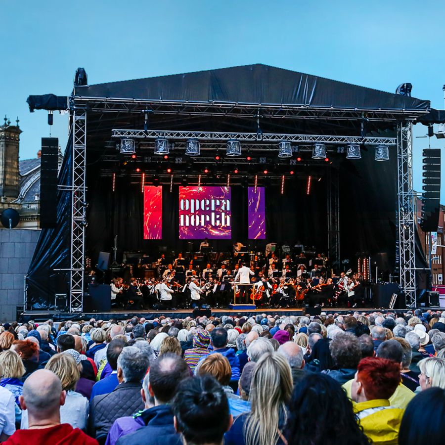 Seated crowd watching the Orchestra of Opera North play on stage at Millennium Square on a bright sunny day