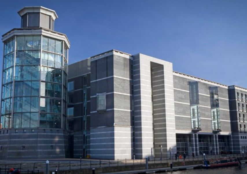 The Royal Armouries Museum, a large modern grey building with a glass tower, sits beside a waterfront under a clear blue sky.