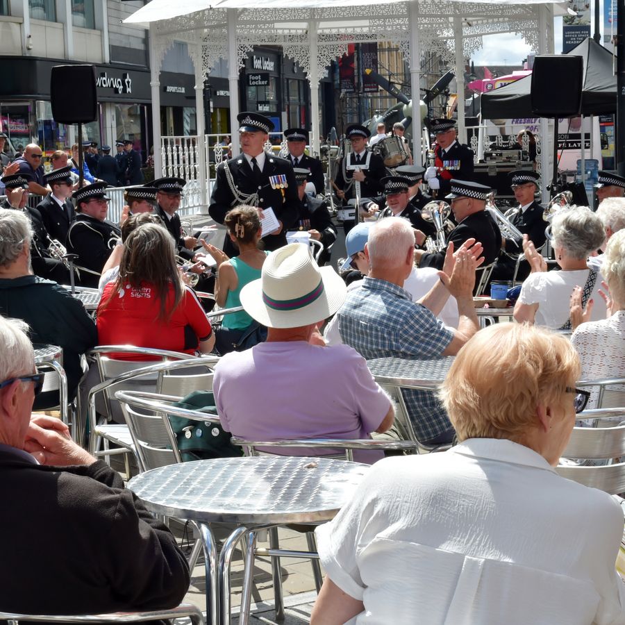 A large crowd sits at outdoor tables on Briggate watching a live performance by a marching band under a white canopy, with shops and city buildings lining the busy pedestrian street on a sunny day.