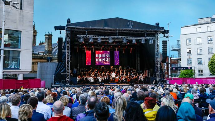 Seated crowd watching the Orchestra of Opera North play on stage at Millennium Square on a bright sunny day