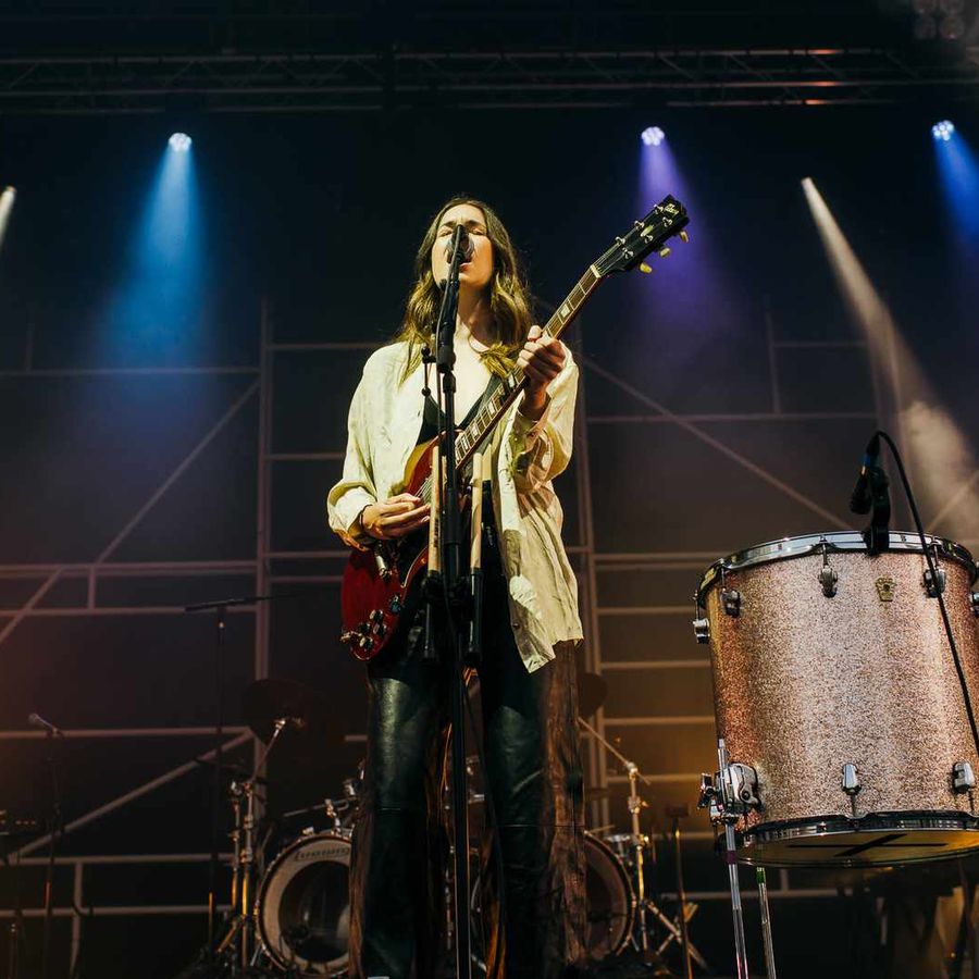Musician playing guitar centre stage with drum kit and band equipment visible behind.