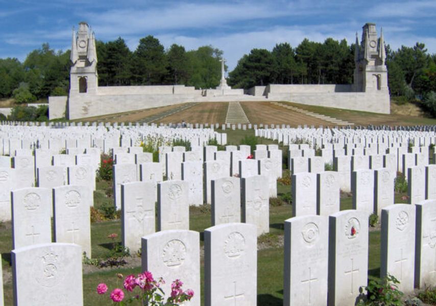 A First World War cemetery with rows of white headstones stretching across a landscaped field, framed by two stone memorial structures and trees in the background.