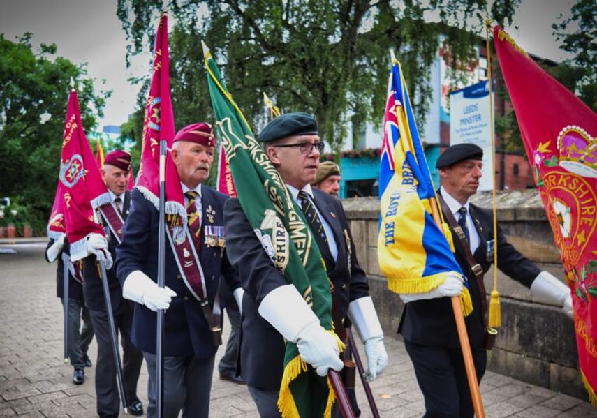 A group of veterans in uniform march in formation carrying colourful regimental standards and flags along a paved walkway in Leeds.