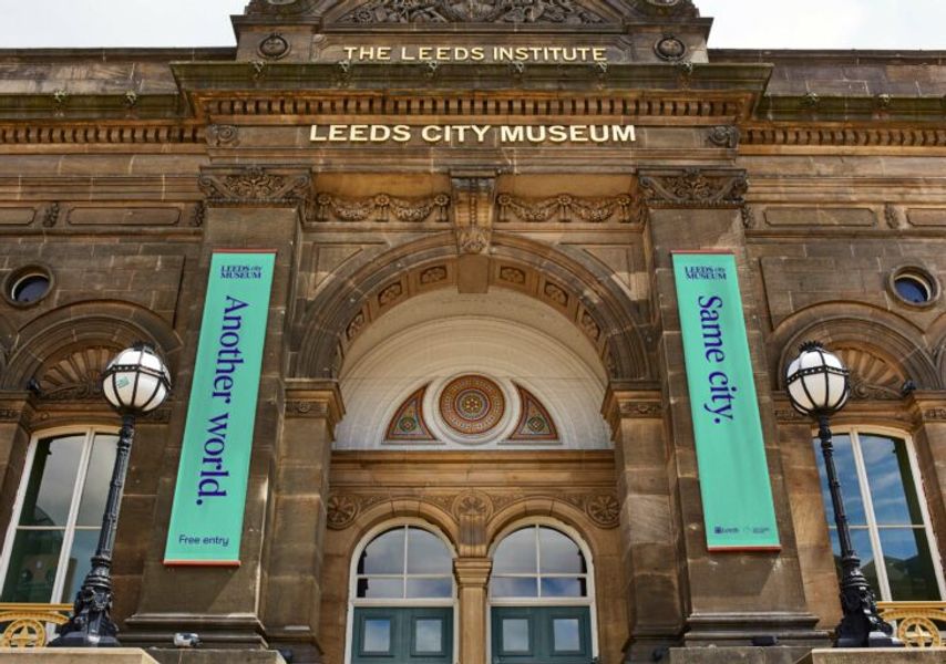 The historic entrance of Leeds City Museum, with stone architecture and banners reading “Another world.” and “Same city.” hanging on either side.